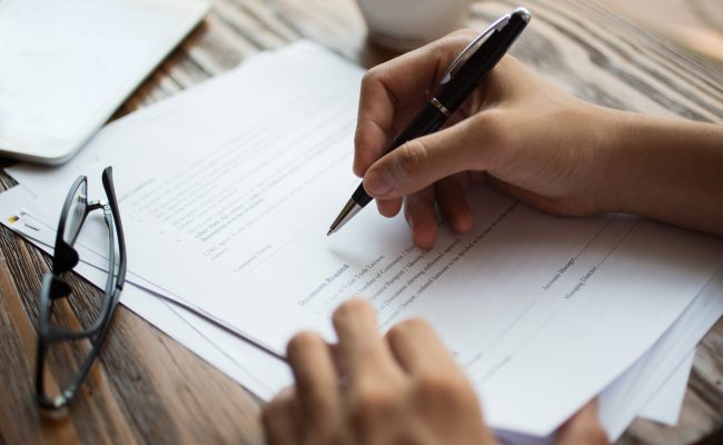 Unrecognizable businessman examining papers at table. Manager with ballpoint pen filling business papers. Close-up of male hands working at desk. Analyzing documents concept